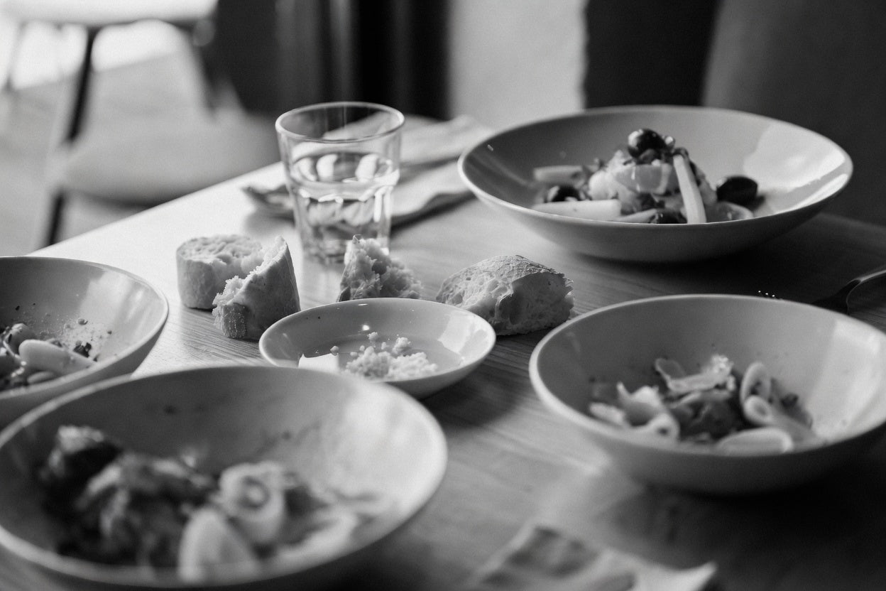 A wooden table after a shared Italian meal — bowls of pasta partially eaten, torn bread, a small dish with crumbs, and a half-full water glass.
