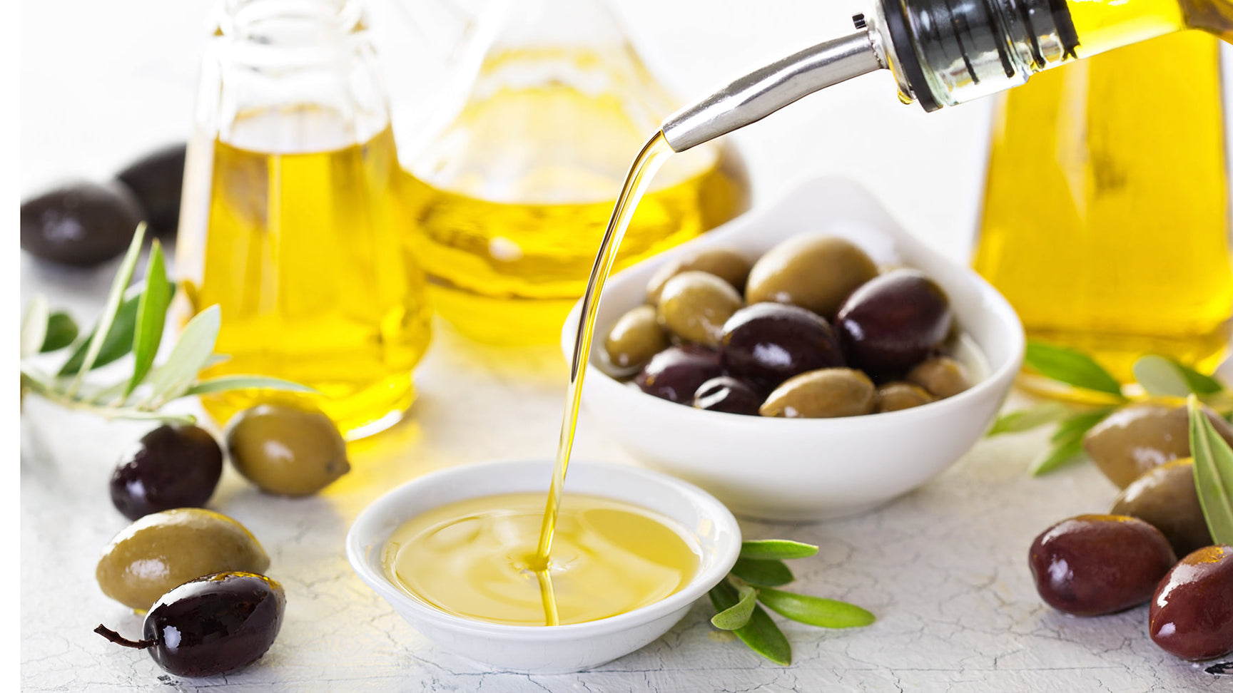 Extra virgin olive oil being poured into a white dish beside green and black olives