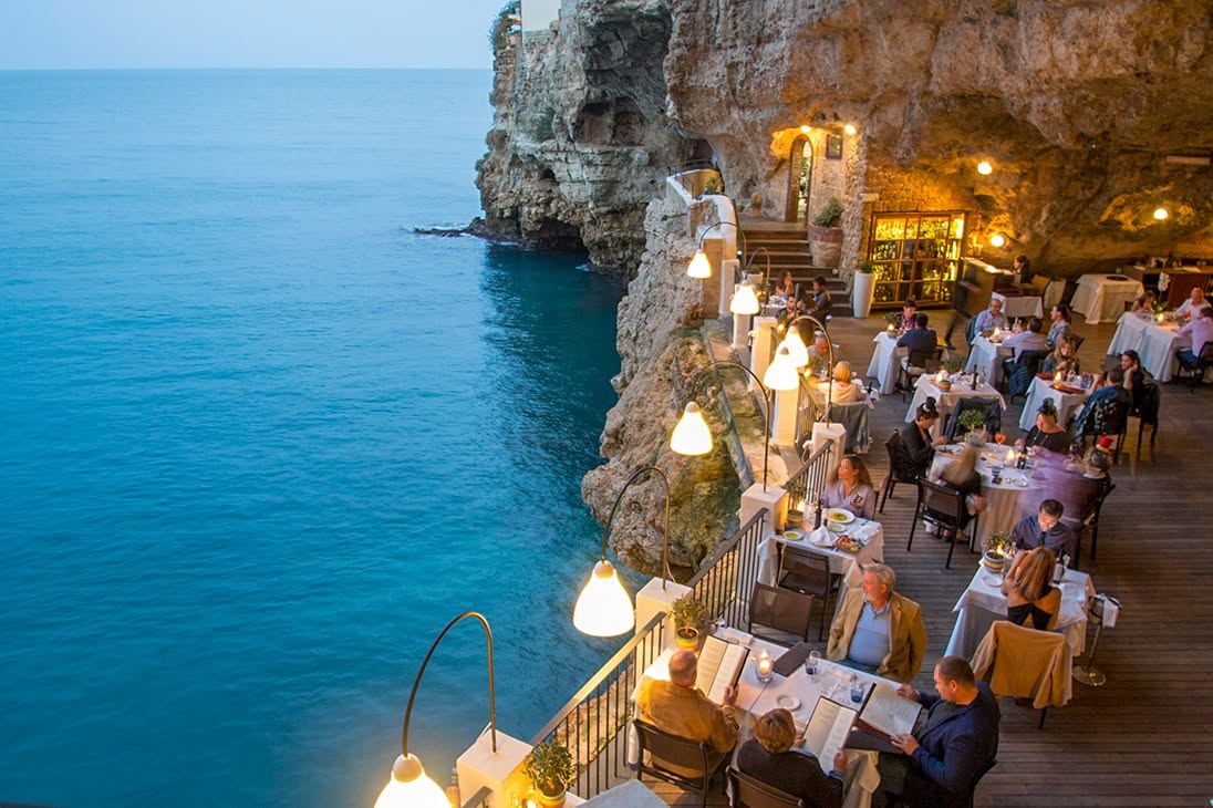 A scenic view of a seaside cliffside restaurant in puglia, italy, with tables and seated diners under warm hanging lights, overlooking bright blue ocean water.