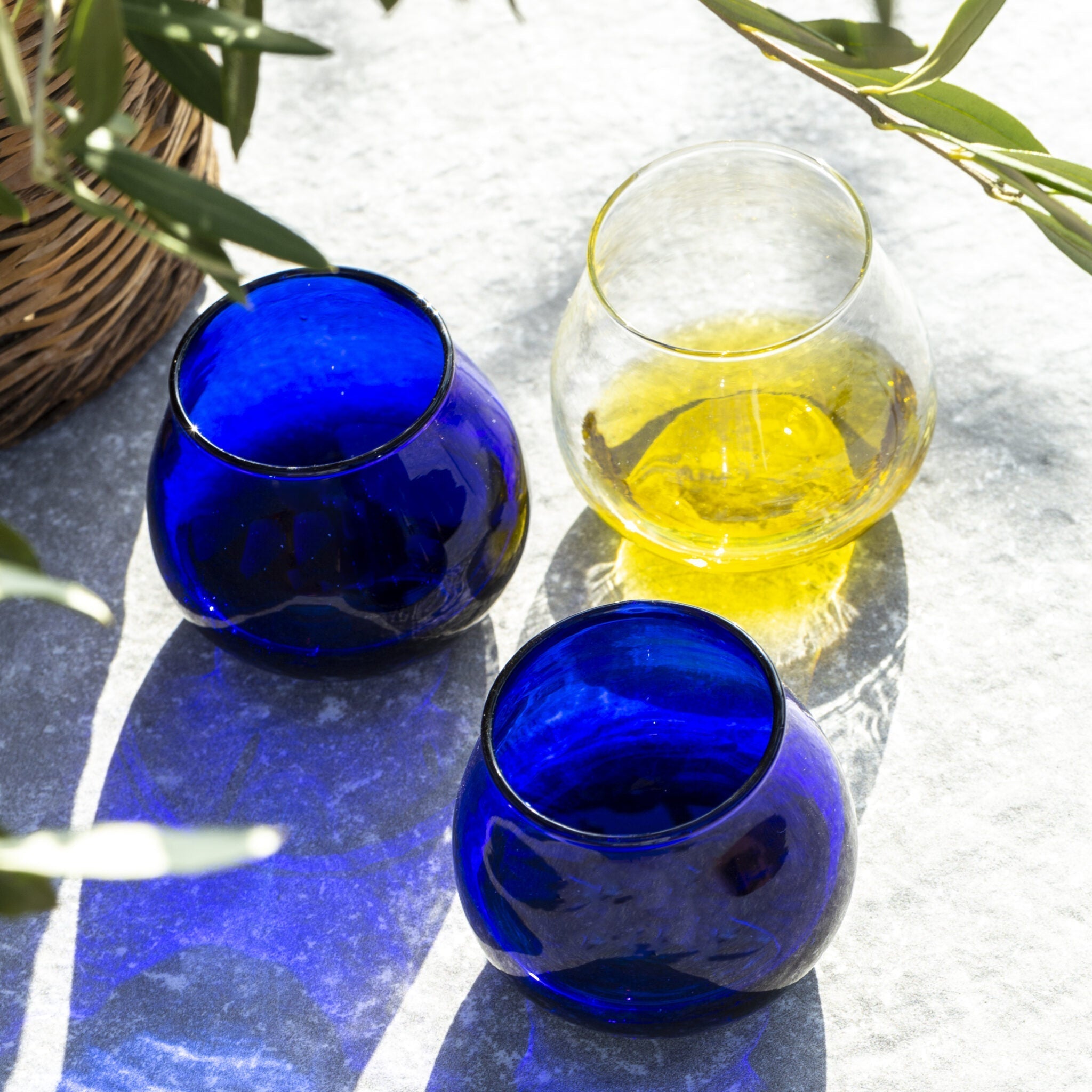 Golden olive oil in a tasting glass beside two blue glasses under olive branches.