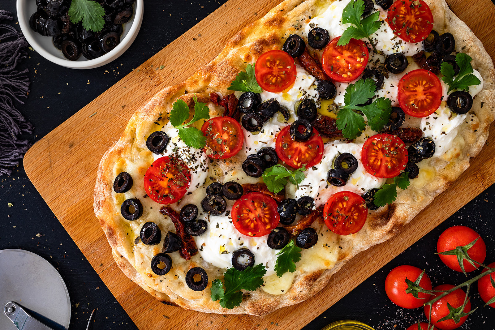 Rectangular pizza topped with cherry tomatoes, black olives, fresh cheese, herbs and olive oil on a wooden board.