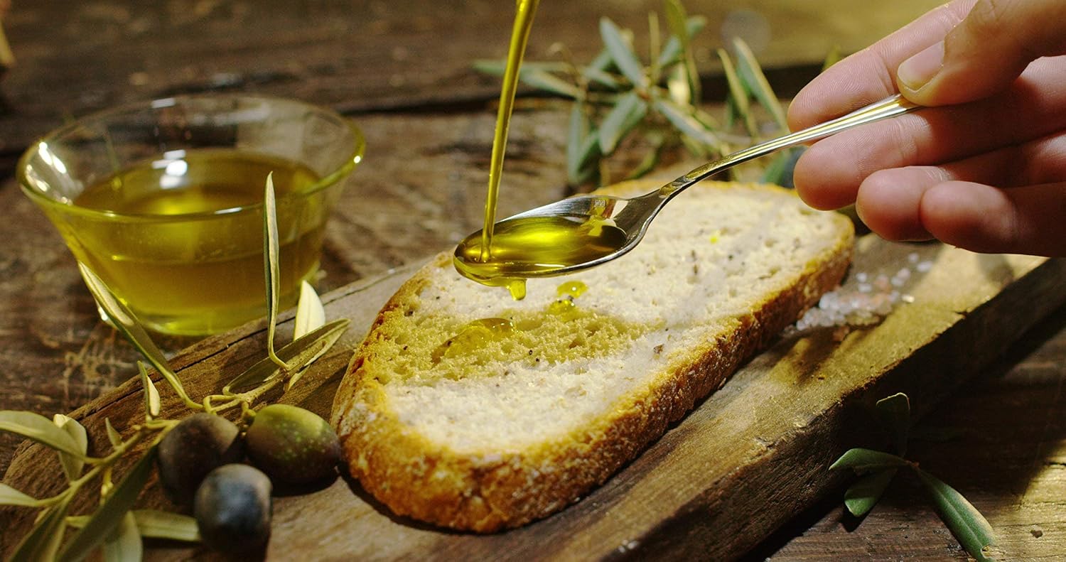 Extra virgin olive oil being spooned onto rustic bread beside fresh olives and a bowl of olive oil.