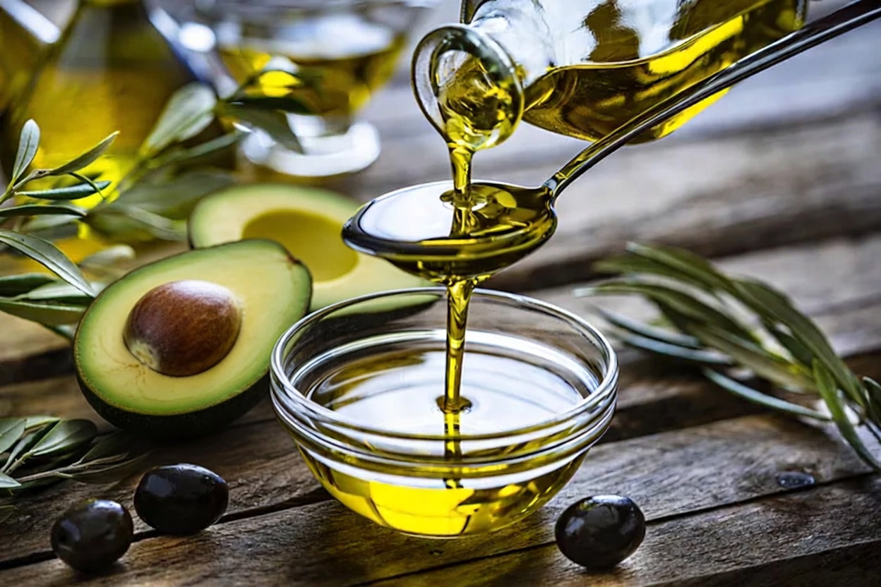 Italian extra virgin olive oil being poured from a glass bottle into a small bowl, beside a halved avocado, fresh olive branches, and black olives on a rustic wooden surface