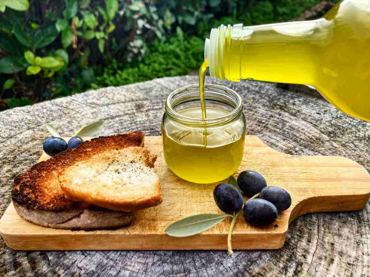 Extra virgin olive oil being poured into a glass jar beside toasted bread and black olives.
