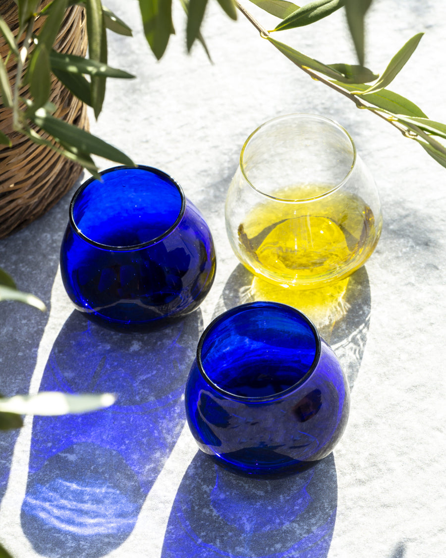 Three stemless oil taste glasses on a light stone surface with olive branches and shadows. Two glasses are cobalt blue, and one is clear glass containing extra virgin olive oil