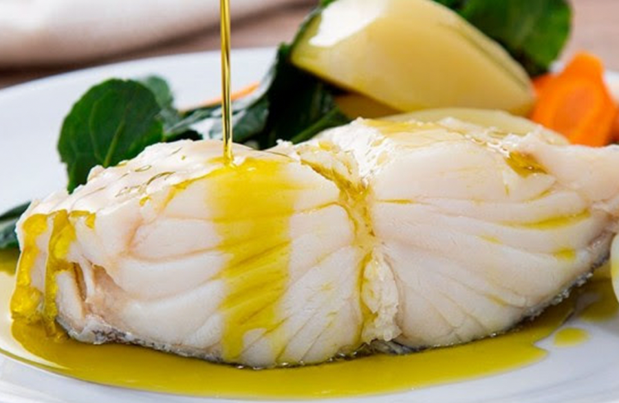A close-up of a cooked fish fillet on a white plate, being drizzled with olive oil, with greens, a lemon wedge, and carrot visible in the background.