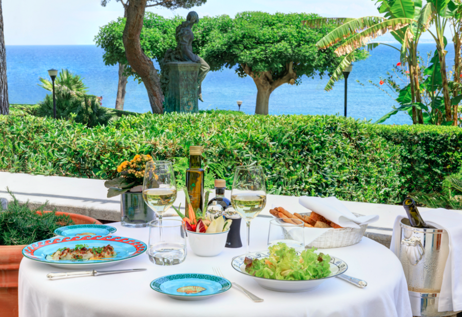 An outdoor mediterranean dining table set with a white tablecloth, plates, wine glasses, bread, and a bottle of olive oil, set against a lush garden with a statue and the blue sea in the background.