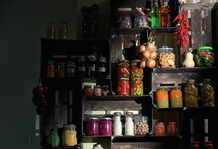 A rustic wooden shelving unit filled with glass jars of pickled vegetables, nuts, spices, and bottles of extra virgin olive oil in a dimly lit kitchen or pantry setting.