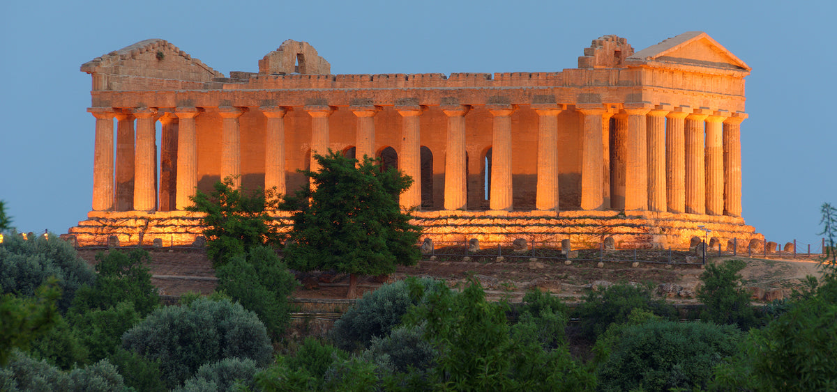 Ancient Greek temple ruins in Agrigento, Sicily, illuminated at dusk beneath a deep blue sky, with green trees in the foreground.
