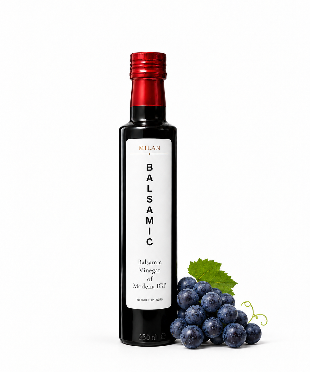 A bottle of Milan Balsamic Vinegar of Modena IGP next to dark grapes and a green leaf on a white background.
