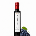 A bottle of Milan Balsamic Vinegar of Modena IGP next to dark grapes and a green leaf on a white background.
