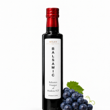 A bottle of Milan Balsamic Vinegar of Modena IGP next to dark grapes and a green leaf on a white background.
